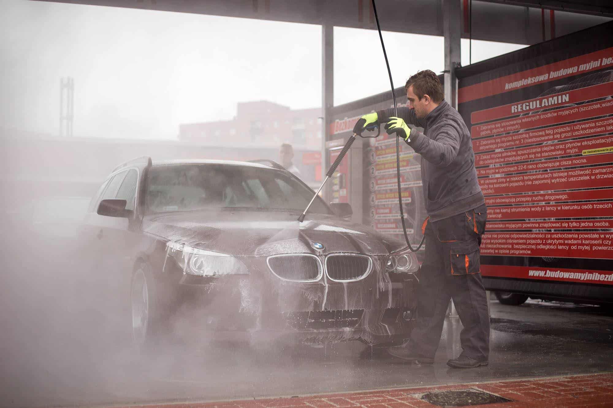 Mężczyzna używa myjki ciśnieniowej do mycia czarnego samochodu w myjni bezdotykowej. W tle widoczne informacje o regulaminie myjni. Proces mycia generuje dużo piany i pary, podkreślając efektywność nowoczesnych myjni bezdotykowych.