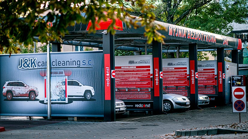 Zewnętrzny widok myjni bezdotykowej z logiem J&K car cleaning, prezentujący strefę mycia samochodów. Na ścianach widoczne informacje dotyczące regulaminu oraz usługi. W tle stoją różne pojazdy, otoczone zielenią drzew.