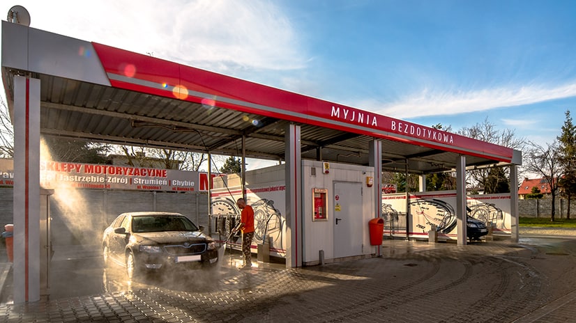 Nowoczesna myjnia bezdotykowa z miejscem do mycia samochodów, otoczona przez urządzenia do czyszczenia. Widoczny samochód pod strumieniem wody, świadczący o skuteczności usług.