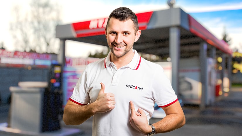 Mężczyzna w białej koszulce z logo "redconst" uśmiecha się i wskazuje na siebie palcami. W tle znajduje się myjnia bezdotykowa, co sugeruje związek z branżą myjni i usług czyszczących.