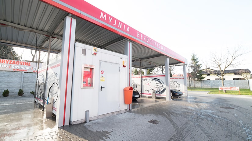 Zewnętrzny widok myjni bezdotykowej z charakterystycznym zadaszeniem i oznaczeniem. Na pierwszym planie widoczne stanowisko mycia, a w tle pojazdy czekające na usługę. Przestrzeń jest czysta i dobrze oświetlona, a otoczenie sprzyja dbaniu o samochód.