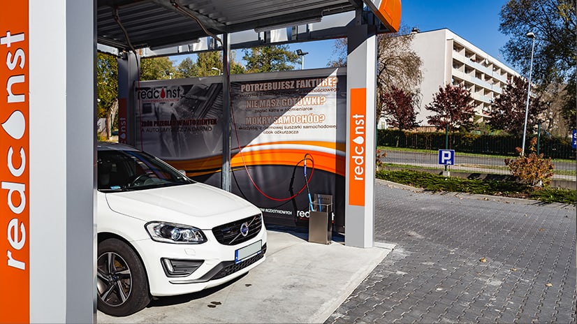 Biała Volvo stoi na myjni bezdotykowej redconst, z reklama na ścianie i wężem do mycia. Idealne miejsce do szybkiego i komfortowego czyszczenia samochodu.