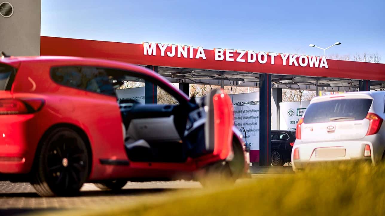 Widok na myjnię bezdotykową z czerwonym samochodem na pierwszym planie oraz innymi pojazdami w tle. W tle widoczny napis „MYJNIA BEZDOTYKOWA” oraz informacje o stacjach diagnostycznych, co ilustruje nowoczesne usługi czyszczenia pojazdów.