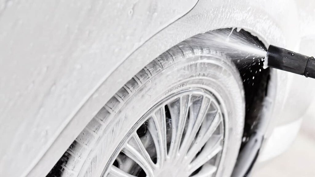 Zbliżenie na koło samochodu w trakcie mycia, z wodą i pianą spływającymi z jego powierzchni. Widoczny proces czyszczenia w myjni bezdotykowej, idealny dla właścicieli pojazdów dbających o ich estetykę.