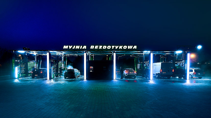 Myjnia bezdotykowa z podświetleniem, gdzie kilka samochodów czeka na czyszczenie. Nocna sceneria, intensywne światła oraz widoczny napis "MYJNIA BEZDOTYKOWA" tworzą nowoczesny wygląd. Idealne miejsce dla właścicieli pojazdów dbających o ich czystość.