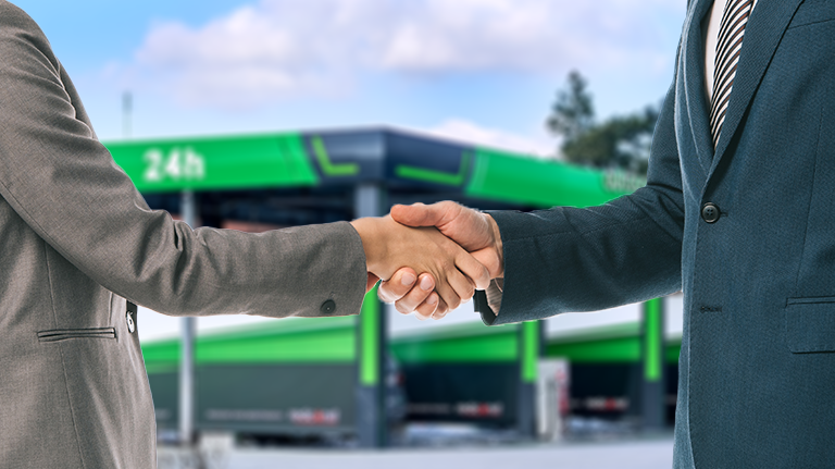 Dwaj mężczyźni w garniturach uścisną ręce na tle nowoczesnej myjni bezdotykowej, co symbolizuje współpracę i partnerstwo w branży motoryzacyjnej.