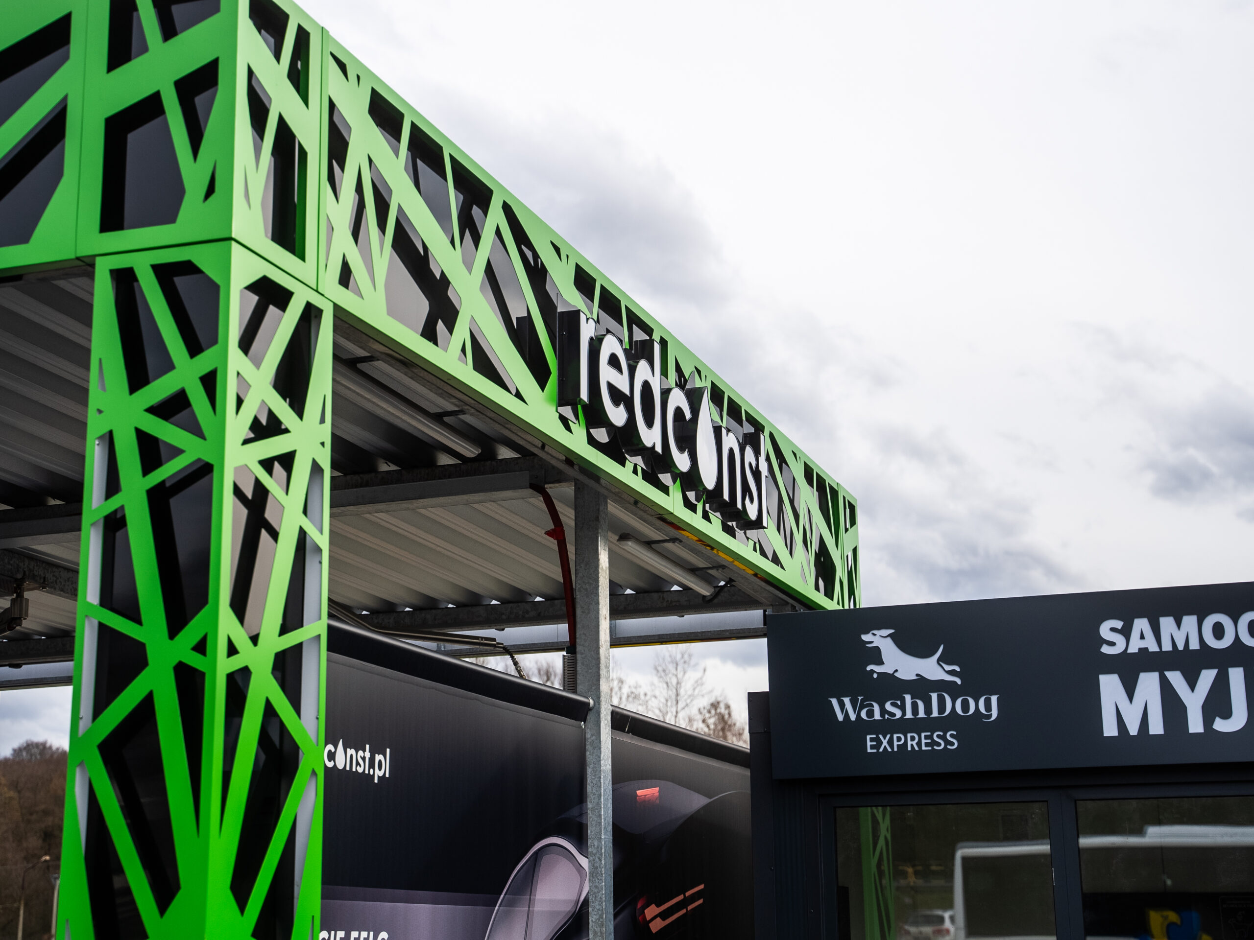 Zielona konstrukcja myjni bezdotykowej z logo "redconst.pl" oraz oznaczeniem "WashDog Express". W tle widoczne chmury, co dodaje dynamiki do nowoczesnej architektury myjni. Idealne dla osób poszukujących efektywnego rozwiązania w myciu pojazdów.