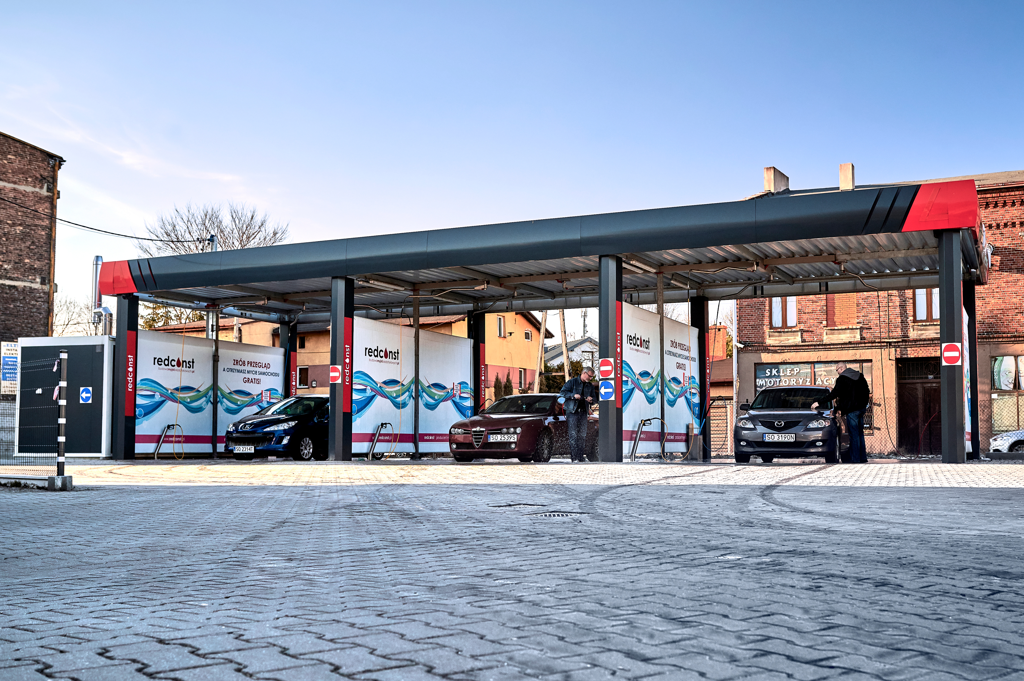 Widok zewnętrzny myjni bezdotykowej, na której dwie osoby myją samochody. W tle kolorowe banery reklamowe oraz budynki. Przestrzeń wygląda nowocześnie i zachęcająco, idealna dla producentów myjni bezdotykowych.