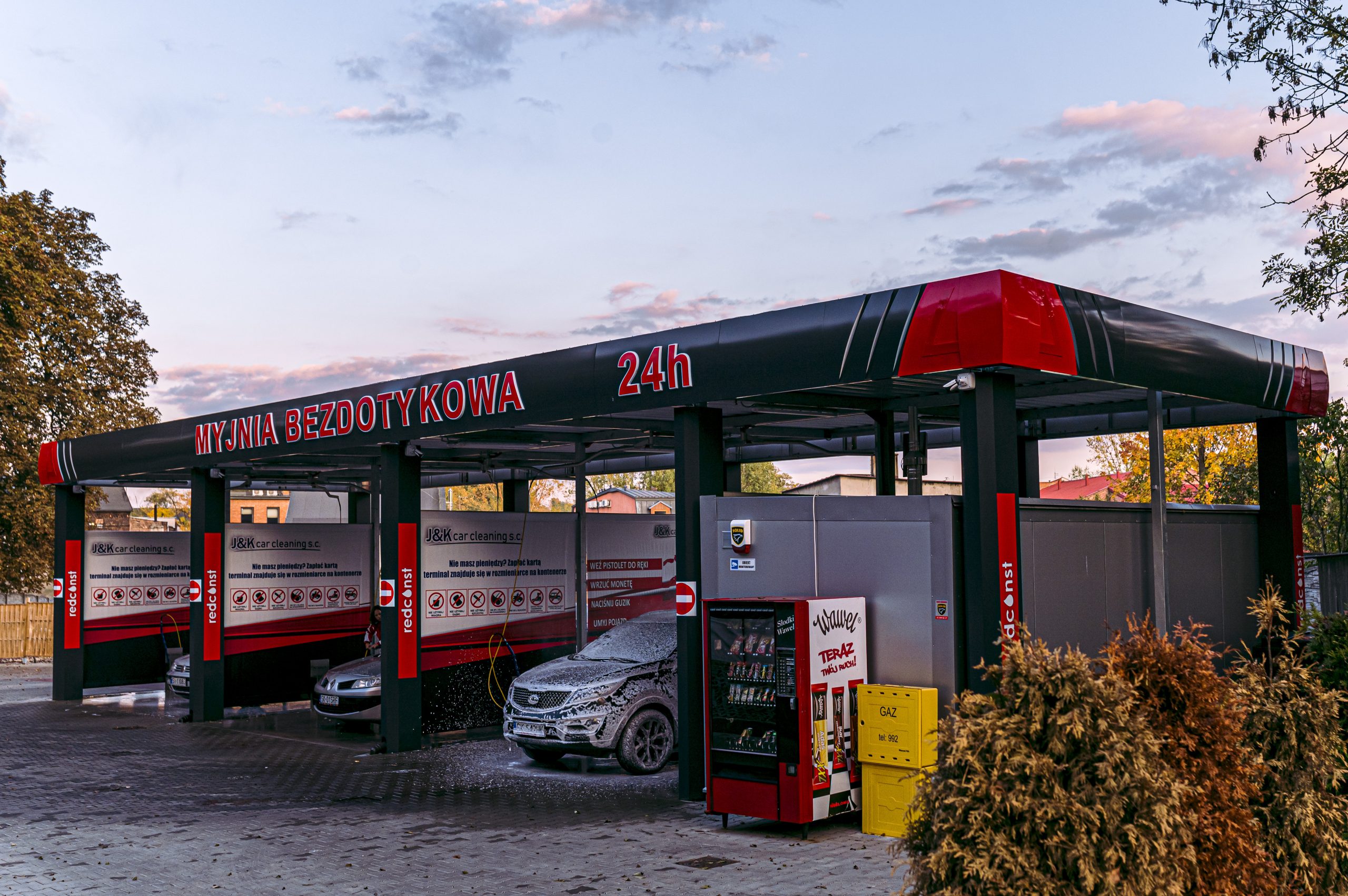 Bezdotykowa myjnia samochodowa otwarta 24 godziny na dobę, z samochodem pokrytym pianą i maszynami do mycia. Dodatkowe elementy, takie jak automat z napojami i informacyjne tablice przy wejściu.