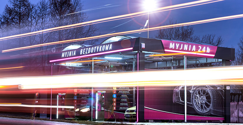 Nowoczesna myjnia bezdotykowa z podświetleniem, otwarta całą dobę, oferująca różne usługi. W tle widać samochody oraz graficzne elementy marketingowe, podkreślające jakość myjni. W nocy dynamiczne efekty świetlne tworzą atrakcyjną atmosferę.