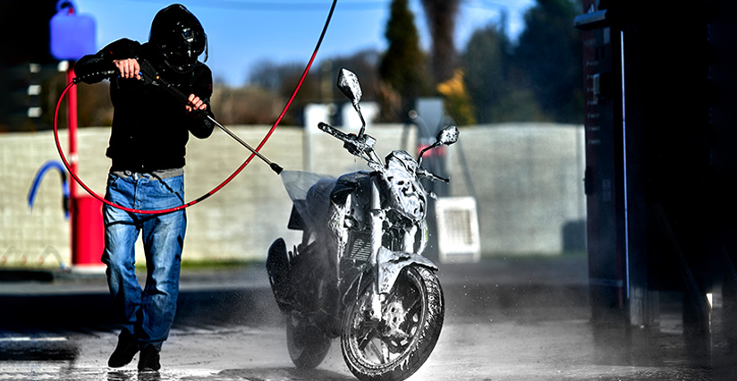 Mężczyzna myje motocykl za pomocą myjki wysokociśnieniowej w myjni bezdotykowej. Woda i piana spływają z pojazdu, a w tle widać infrastrukturę myjni, co podkreśla efektywność mycia motocykli.