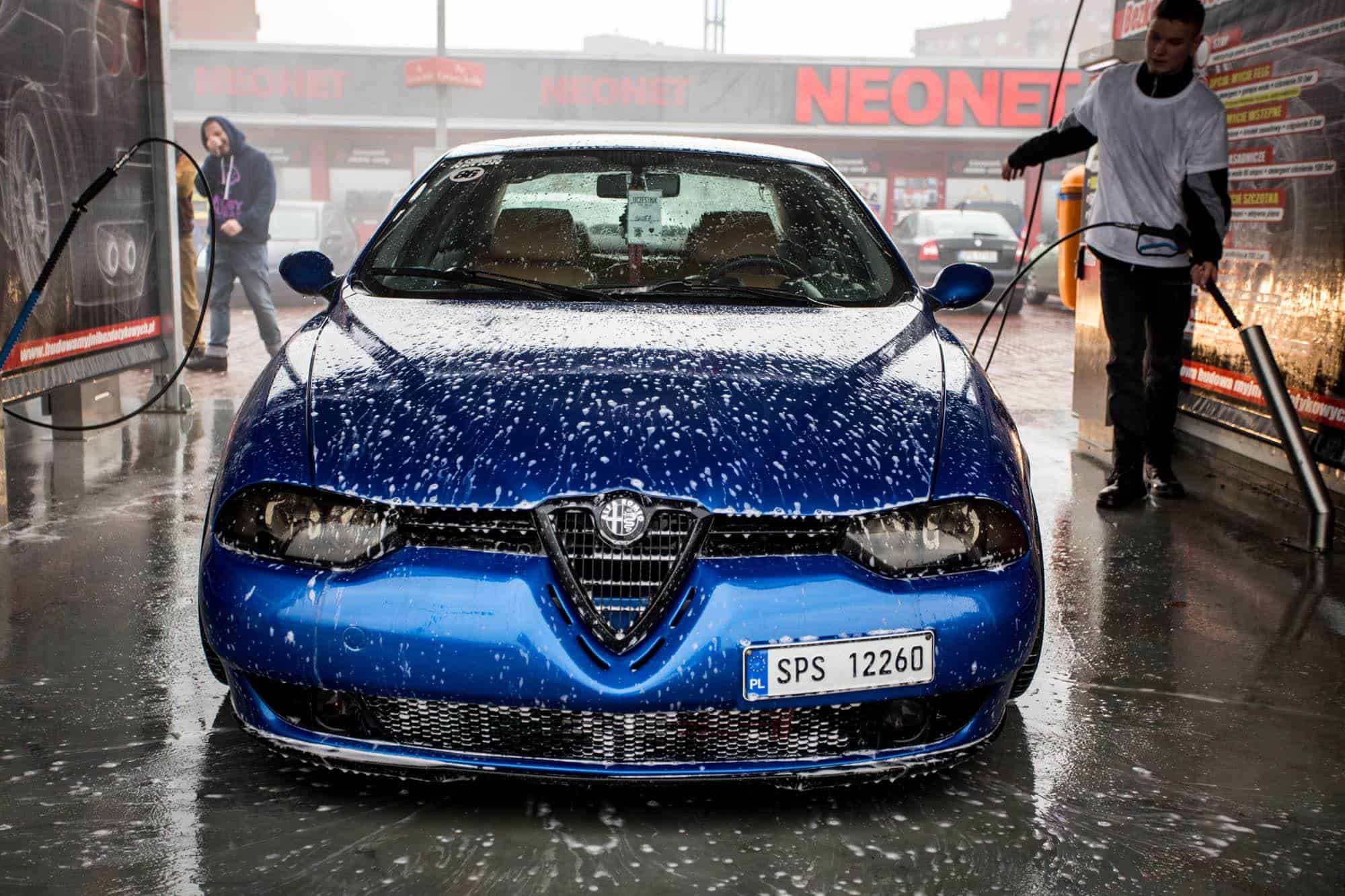 Niebieskie auto myte w myjni bezdotykowej, podczas aplikacji piany przez pracownika. Tło z logo "NEONET" i innymi pojazdami na parkingu, tworzy dynamiczną scenerię myjni. Idealne zdjęcie ilustrujące nowoczesne myjnie bezdotykowe.