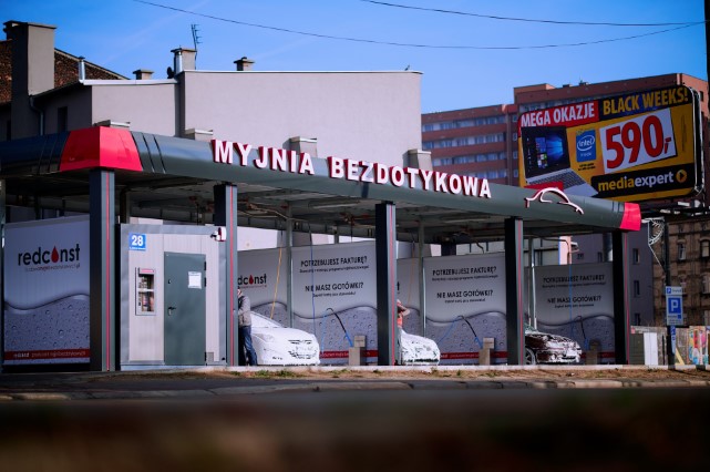 Widok myjni bezdotykowej z napisem "MYJNIA BEZDOTYKOWA", z samochodami przygotowanymi do mycia. W tle reklama z promocjami. Zdjęcie ilustruje nowoczesne usługi czyszczenia pojazdów w mieście.
