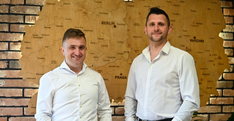 Dwóch mężczyzn w białych koszulach, uśmiechających się przed mapą przedstawiającą Europę Środkową, ze szczególnym uwzględnieniem Berlina, Warszawy i Pragi, co może sugerować działalność gospodarczą w regionie, w tym branżę myjni bezdotykowych.