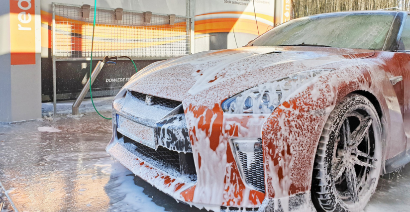 Samochód sportowy pokryty gęstą pianą w myjni bezdotykowej. W tle widoczna infrastruktura myjni oraz wąż do spłukiwania. Idealny przykład nowoczesnego procesu mycia pojazdów.