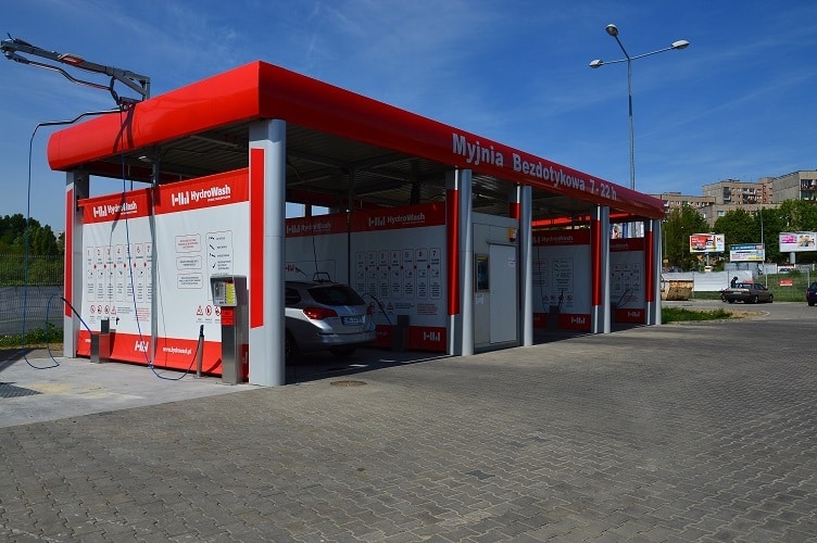 Nowoczesna myjnia bezdotykowa z czerwonym dachem, oferująca usługi mycia samochodów. W tle widać instrukcje obsługi, a na placu stoją pojazdy. Idealne miejsce dla kierowców szukających skutecznego czyszczenia bezdotykowego.