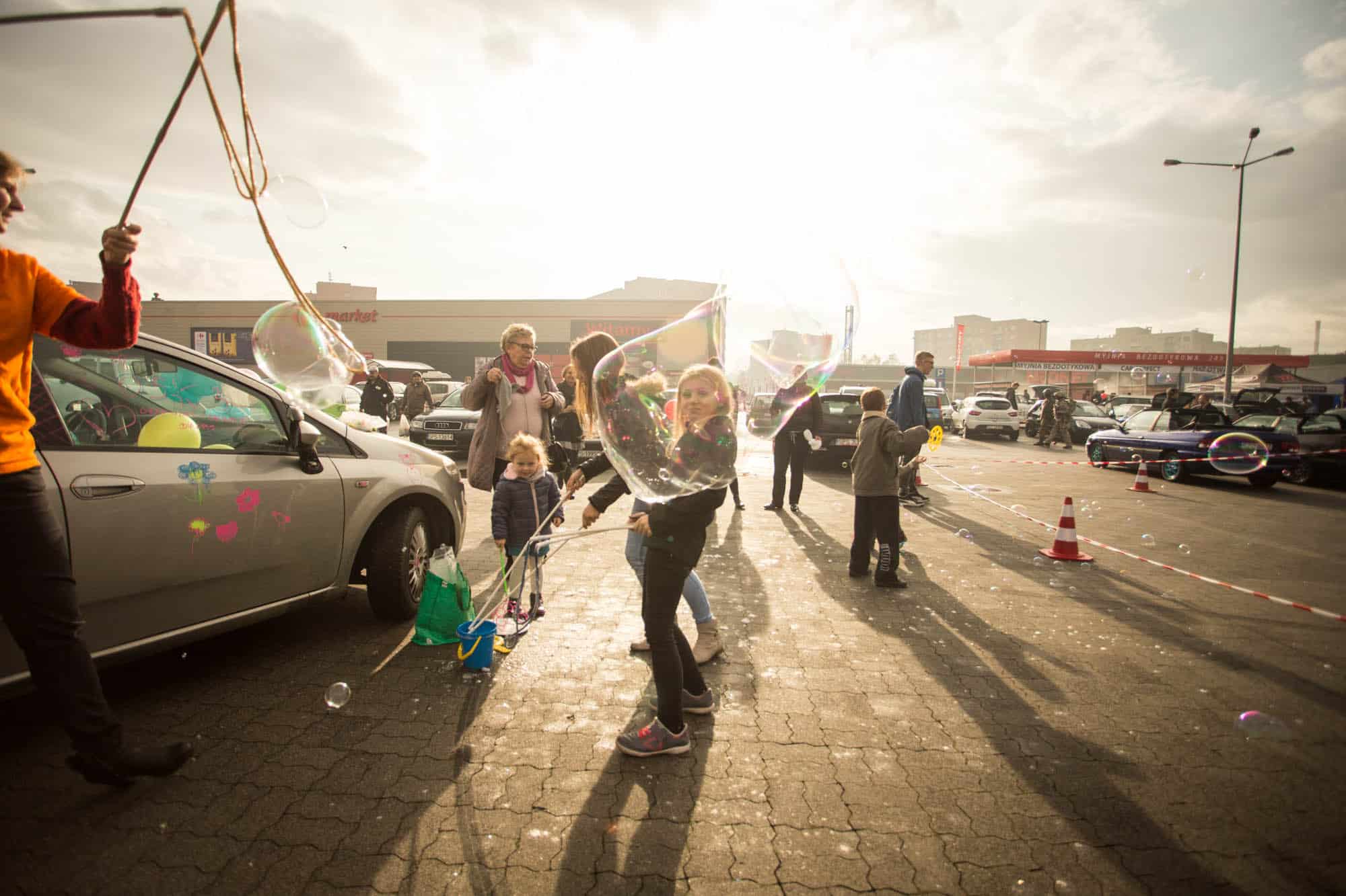 Dzieci bawią się na parkingu, tworząc bańki mydlane. W tle znajdują się samochody, a w powietrzu unosi się słoneczne światło. Scena pełna radości i zabawy, idealna dla rodziny. Event organizowany przez producenta myjni bezdotykowej.