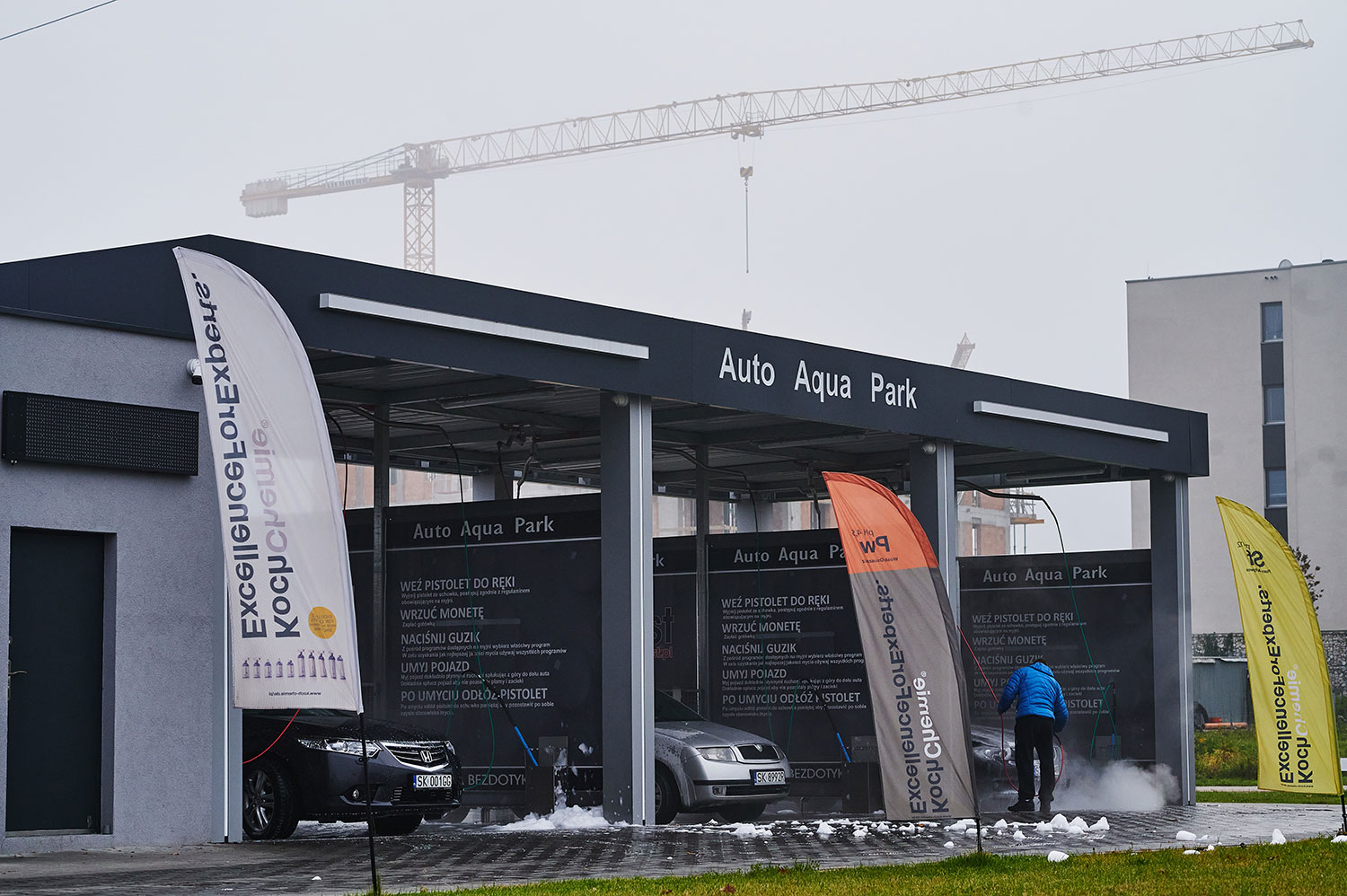 Nowa myjnia bezdotykowa Auto Aqua Park z flagami reklamowymi, samochody w trakcie mycia. W tle widoczny dźwig budowlany, co sugeruje rozwój okolicy. Idealne miejsce dla właścicieli samochodów, którzy cenią sobie wygodę i skuteczność mycia.