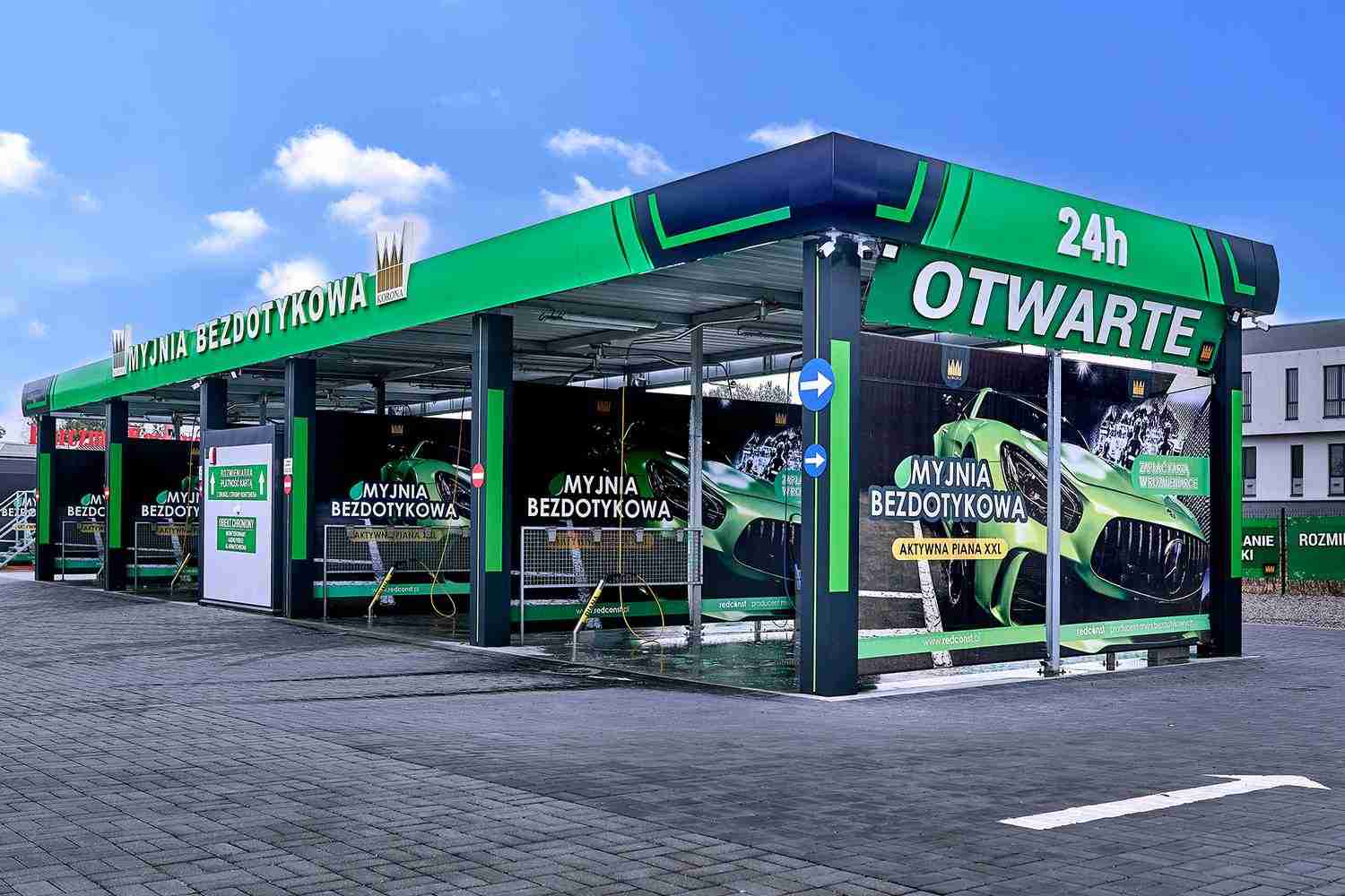 Nowoczesna myjnia bezdotykowa, otwarta 24 godziny na dobę. Zielona konstrukcja z efektywnymi stanowiskami do mycia samochodów, oferująca wyspecjalizowane usługi czyszczenia. Dodatkowe informacje na reklamach umieszczonych na budynku myjni. Idealna dla właścicieli pojazdów poszukujących wygody.