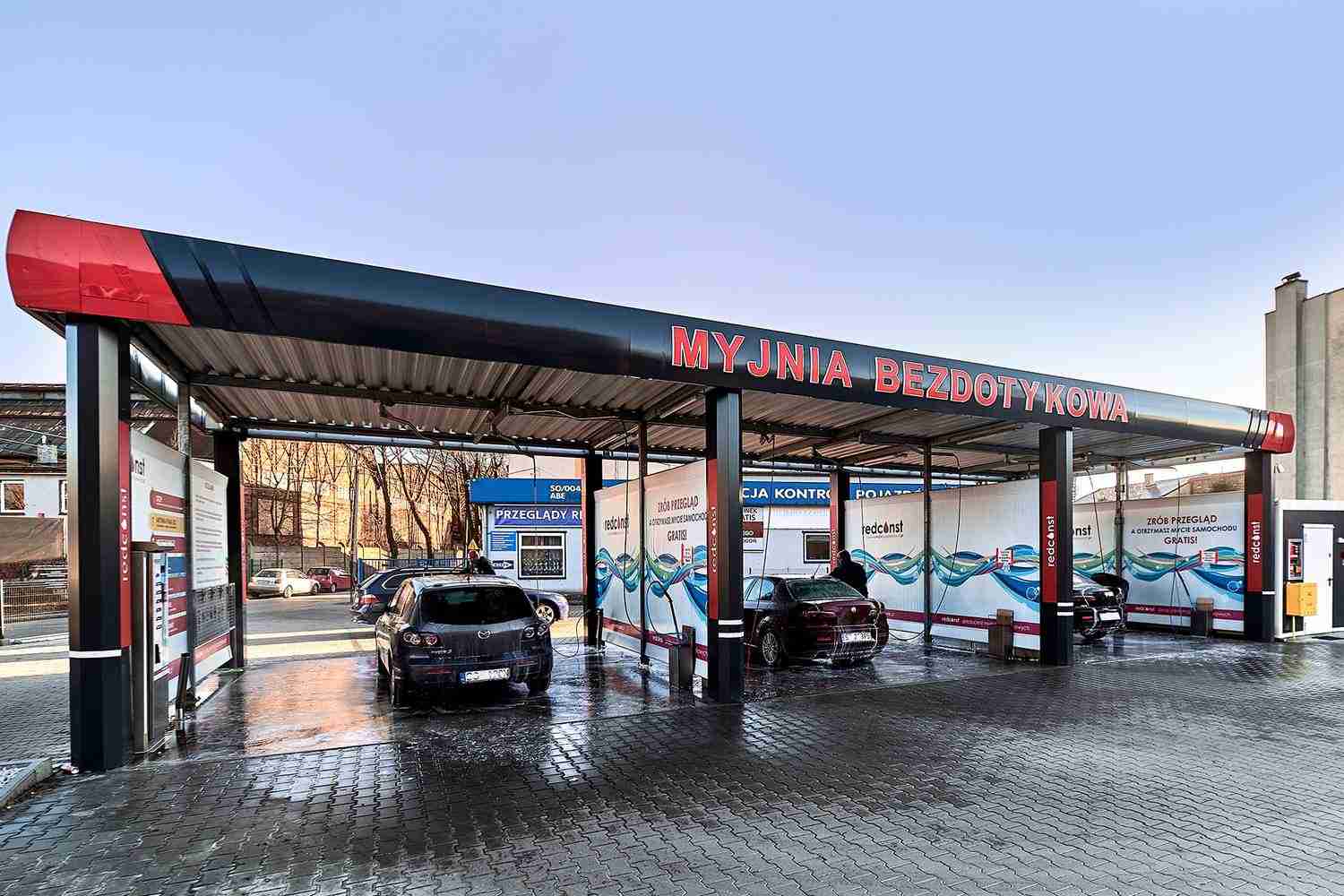 Nowoczesna myjnia bezdotykowa z dwoma samochodami w trakcie czyszczenia. Wyraźne oznaczenia oraz infrastruktura sprzyjająca efektywnemu myciu pojazdów. Czyste, wykafelkowane powierzchnie i jasne niebo w tle podkreślają innowacyjny charakter miejsca.