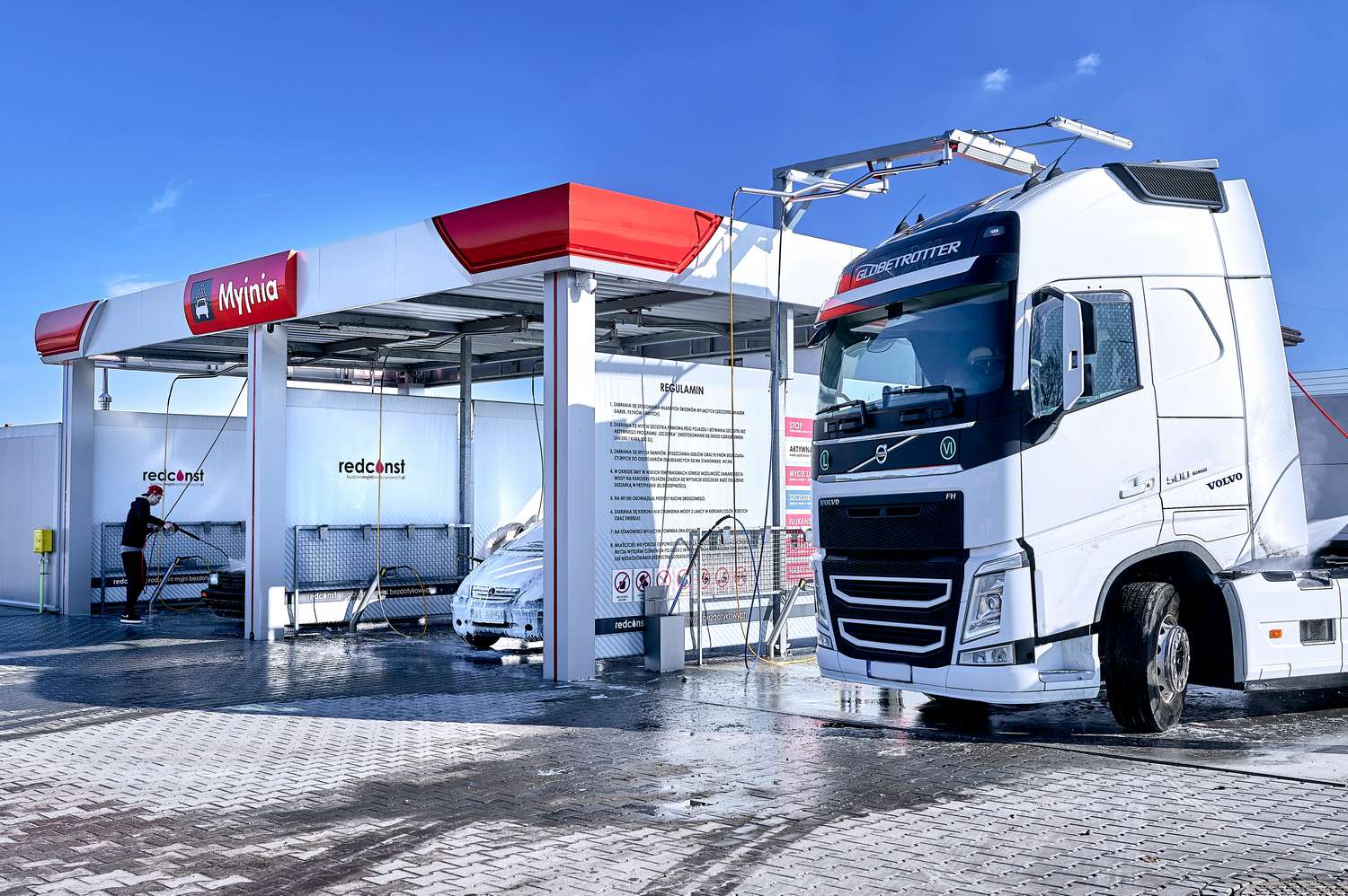 Myjnia bezdotykowa z dużym ciężarowym pojazdem Volvo w trakcie mycia. W tle widoczne strefy do mycia z wężami i instrukcją obsługi. Słoneczne niebo oraz mokra powierzchnia myjni podkreślają nowoczesny wygląd obiektu.