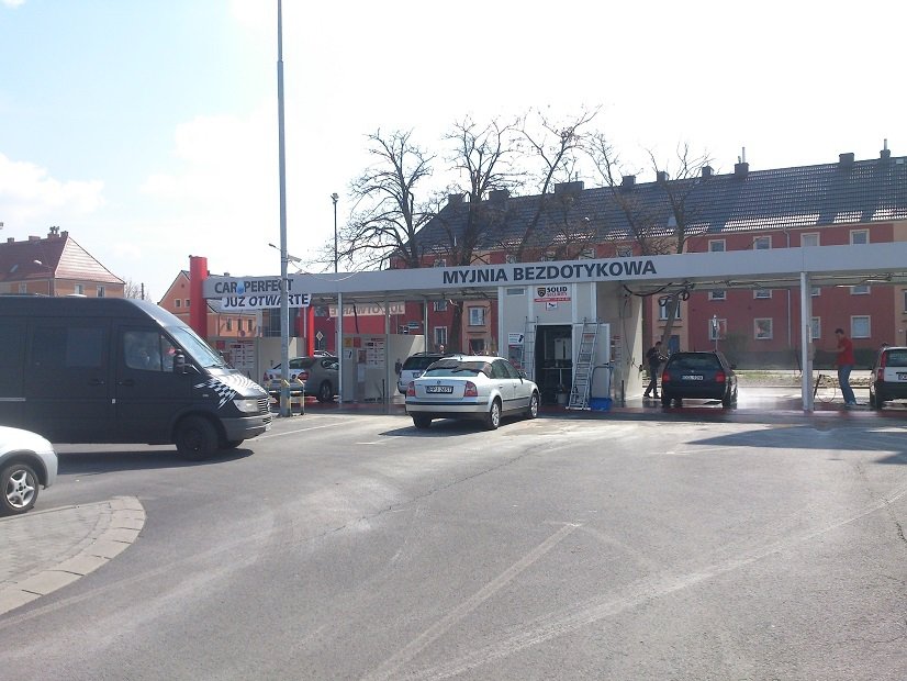 Widok na myjnię bezdotykową, gdzie kilka samochodów korzysta z usług. Przed obiektem widoczny jest znak informujący o otwarciu, a w tle znajdują się mieszkalne budynki. Myjnia to idealne miejsce dla kierowców dbających o czystość swoich pojazdów.