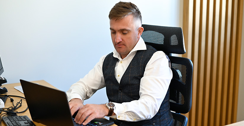 Mężczyzna w eleganckim stroju siedzi przy biurku, korzystając z laptopa. W tle widoczne elementy nowoczesnego biura. Idealne dla producentów myjni bezdotykowych, ilustracja profesjonalnego środowiska pracy.