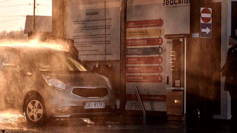 Zbliżenie na samochód myty w myjni bezdotykowej, gdzie strumień wody i piany pokrywa pojazd. W tle widoczny panel z instrukcjami oraz numeracja mycia. Słoneczna aura nadaje scenie ciepły klimat, podkreślając funkcjonalność myjni. Idealny obraz producenta myjni bezdotykowych.