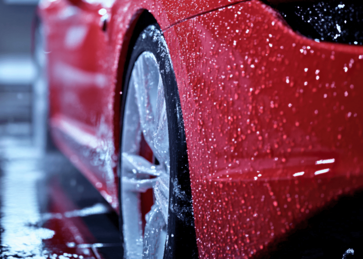 Zbliżenie na czerwony samochód myty w myjni, z kropelkami wody i piany na karoserii i oponach, ukazujące efekty mycia bezdotykowego. Idealny obrazek dla producentów myjni bezdotykowych promujących usługi czyszczenia pojazdów.